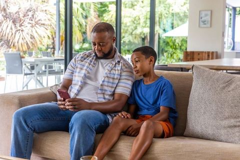 African american father and son sharing smartphone on sofa, bonding in bright modern home