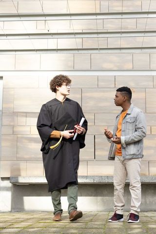 Graduate walking and chatting with friend on sunlit campus plaza holding diploma