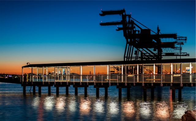 Industrial pier with structural elements silhouetted against the vibrant sunset, creating a striking visual with reflections on the water. Suitable for use in topics related to engineering, architecture, urban landscapes, or evening waterfront tranquility.