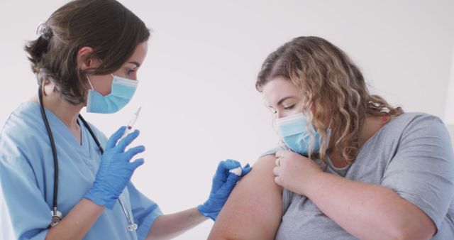 Nurse wearing blue scrubs and mask administering COVID-19 vaccine to woman in protective face mask, underscoring importance of vaccination for public health; suitable for illustrating healthcare providers, vaccination programs, and COVID-19-related content.