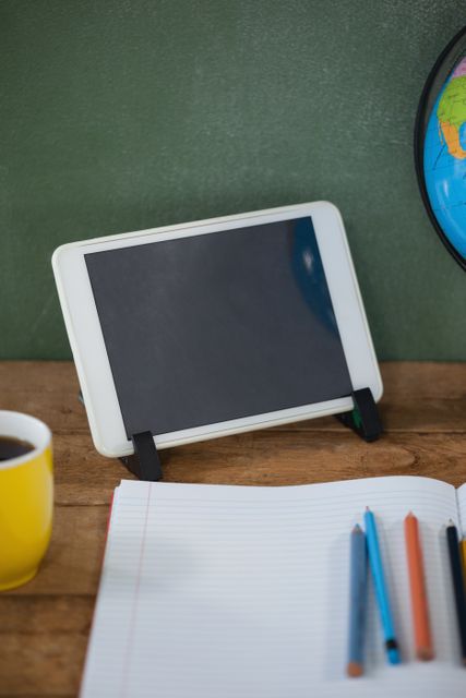 Digital tablet and globe on a wooden desk in a classroom setting. Notebook, pencils, and coffee cup also visible. Ideal for educational content, school-related articles, and technology in education themes.