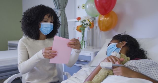 Mother is sitting by her daughter's hospital bed reading a card. The daughter is lying in bed holding a teddy bear while both are wearing surgical masks. Balloons and flowers are seen in the room. This can be used in healthcare promotions, family support articles, or materials related to hospital care and COVID-19 precautions.