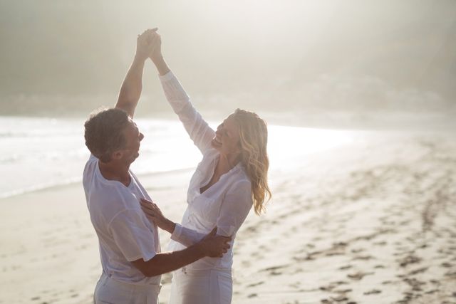 This image depicts a happy mature couple dressed in white clothing, enjoying a sunny day at the beach. They are holding hands, smiling, and looking at each other, expressing joy and togetherness. Perfect for use in advertisements or marketing materials promoting travel, vacations, romantic getaways, retirement, relationships, or active lifestyles for older adults.