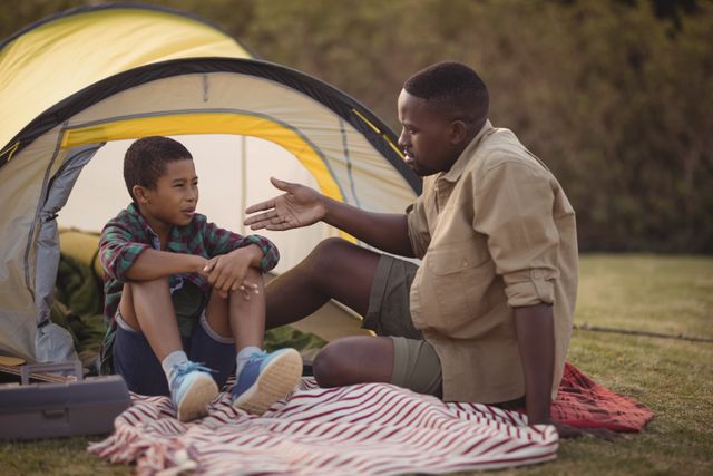 Father comforting his son outside a tent in a park on a sunny day. This image can be used for themes related to family bonding, parenting, outdoor activities, emotional support, and leisure time. Ideal for articles, blogs, and advertisements focusing on family relationships, camping trips, and fatherhood.