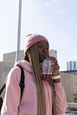 African american woman sipping berry drink in pink beanie and hoodie on city street