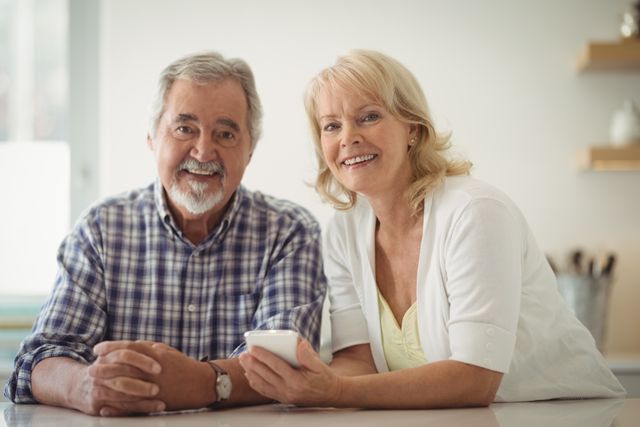 Senior couple smiling and using a mobile phone in a bright kitchen. Ideal for themes related to senior lifestyle, technology use among elderly, family bonding, and retirement living.