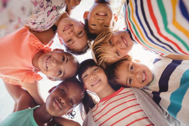 Group of diverse schoolchildren forming a huddle and smiling on a sunny day. Ideal for promoting teamwork, diversity, childhood, education, and outdoor activities. Can be used in educational materials, advertisements, and community-building campaigns.