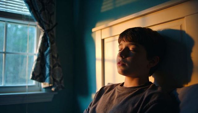 Sunlit teen resting against headboard with window shadows, quiet evening mood