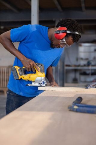 Carpenter cutting wooden plank with cordless jigsaw, wearing ear protection and safety goggles