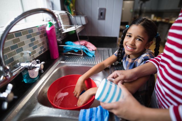 This image shows a young girl smiling while helping her mother wash dishes in the kitchen. It captures a moment of family bonding and teamwork in a domestic setting. Ideal for use in articles or advertisements about family life, parenting, household chores, and teaching children responsibility.