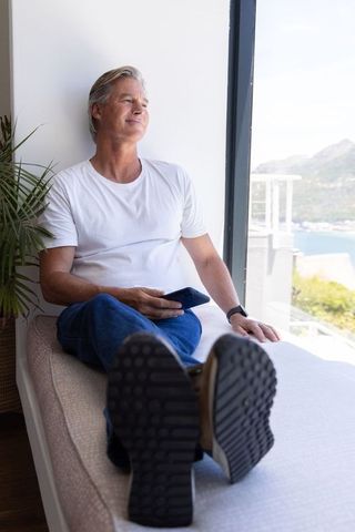 Mature man relaxing on daybed by window holding smartphone and gazing at coastal view