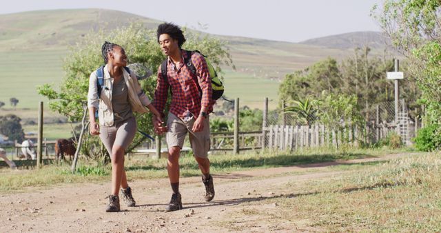 A young couple is hiking together on a countryside path surrounded by lush greenery and hilly landscape. They are carrying backpacks and wearing casual outdoor clothing, smiling and enjoying the sunny day. This image can be used for travel blogs, outdoor adventure websites, nature conservation campaigns, and leisure activity promotions.