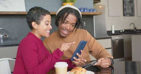 Young diverse couple sharing smartphone at cozy coffeehouse with pastry and tablet
