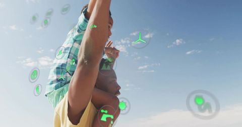 Black father carrying son on shoulders under blue sky with eco ar icons, family bonding