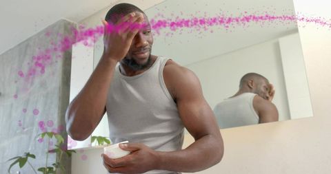 Muscular man applying moisturizer at bathroom mirror holding jar during morning grooming