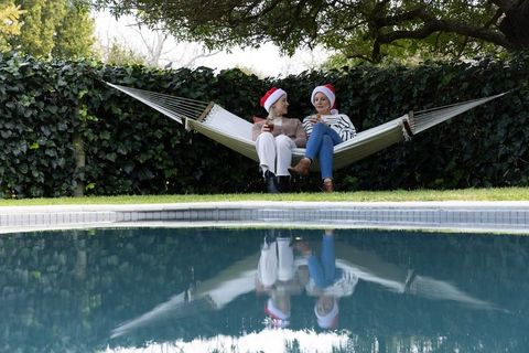Friends wearing santa hats sitting on hammock by backyard pool with water reflection