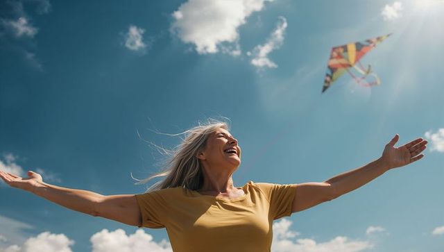 Joyful senior woman stretching arms under blue sky with colorful kite flying overhead