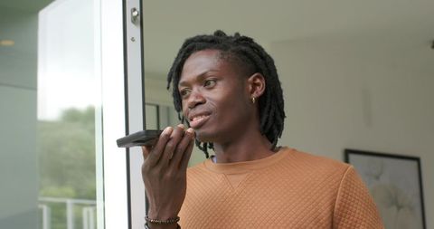 African American man speaking into smartphone while standing by glass door in modern home