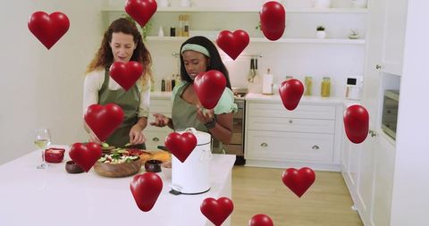 Two women preparing salad together in bright modern kitchen with floating hearts