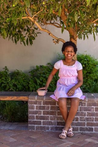 African american girl sitting smiling on ledge in sunlit courtyard wearing pink dress