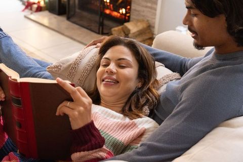 Couple cuddling on sofa by fireplace reading red-bound book wearing cozy knit sweaters