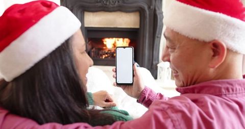Senior asian couple wearing santa hats sharing smartphone by fireplace, cozy holiday joy