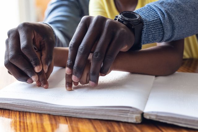 African american father holding hands of daughter and assisting her in reading braille book at home. Unaltered, family, togetherness, childhood, learning, education and disability concept.
