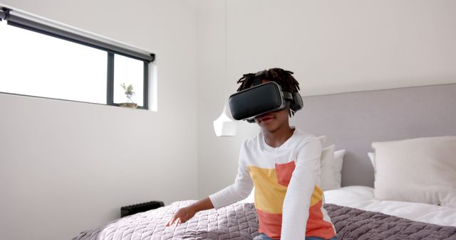 A young child is sitting on a bed in a modern, minimalistic bedroom while wearing a virtual reality headset, appearing engaged and fascinated. This image can be used in technology-related content, articles about VR use among youth, or advertisements for VR headsets and tech gadgets for children.