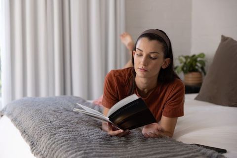 Woman reading paperback on bed, relaxing morning in minimal neutral bedroom, calm lifestyle
