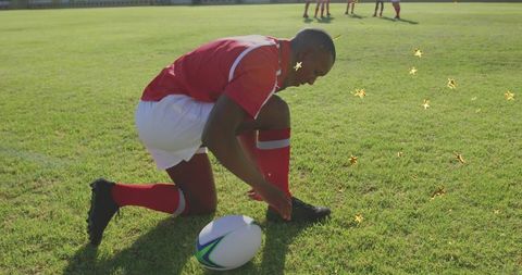 Kneeling rugby player tying cleats on sunny grass pitch with rugby ball nearby