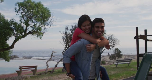 Young couple enjoying a playful piggyback ride near the ocean on a sunny day. They are smiling and having fun, showcasing joy and togetherness. Useful for marketing materials on travel, relationship-building, outdoor activities, and happiness.