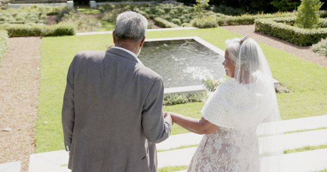 Rear view of happy diverse senior bride and groom holding hands on wedding day in sunny garden. Summer, marriage, romance, love, celebration, tradition and senior lifestyle, unaltered.