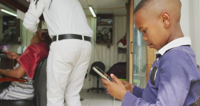 Young boy engaging with a smartphone while seated at a barber shop. Feelings of calmness, distraction, and modernity depicted, perfect for content on children using technology, everyday life, and grooming salons.