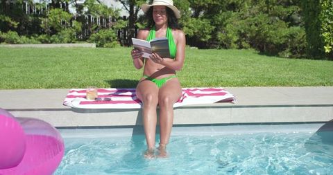African american woman reading by pool in green bikini and sun hat, summer relaxation