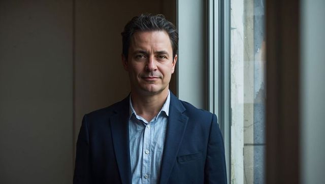 Confident businessman standing by window in office wearing navy blazer and checked shirt