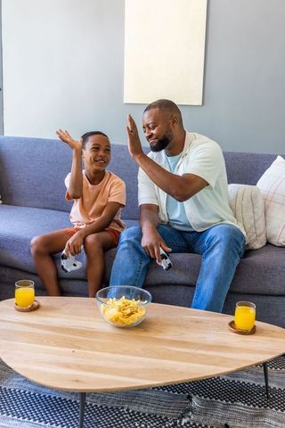 African american father and son playing video games on sofa sharing snacks exchanging high five