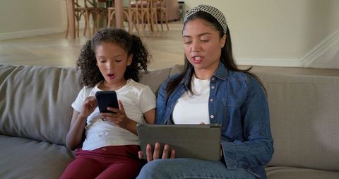 Mother and daughter bonding on sofa, reading tablet and phone together for family learning