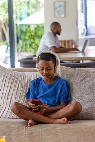 African american dad working remotely while son listening to music using smartphone on couch