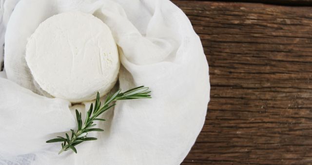 Fresh homemade cheese wheel wrapped in light cloth placed on wooden surface with rosemary garnish. Suitable for illustrating artisanal cheese making, farm-to-table concepts, rustic food presentations, and organic food products. Ideal for food blogs, culinary websites, and advertisements promoting fresh and natural foods.