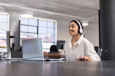 Smiling Businesswoman Wearing Headphones Using Dual Screens in Office