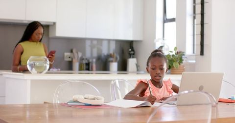 African mother checking phone while daughter studying with laptop at modern kitchen table