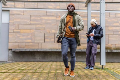 Man walking in tan beanie and green scarf holding smartphone and carrying crossbody bag