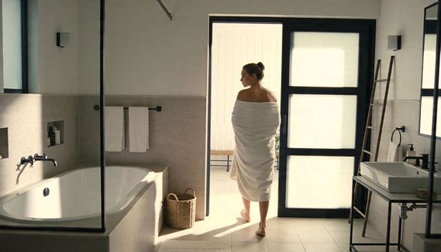 Woman wrapped in towel walking toward frosted glass door in modern minimalist bathroom