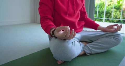 Man meditating cross-legged in red hoodie on green yoga mat by window for mindfulness