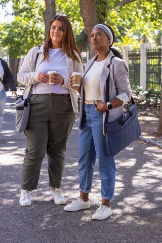Friends enjoying coffee walk on tree-lined city sidewalk, chatting and carrying shoulder bags