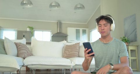 Young asian man listening to music with headphones and using smartphone in bright living room