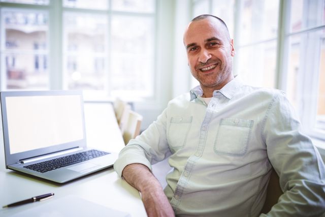 Businessman sitting by laptop in bright office, smiling confidently. Ideal for use in business, technology, and professional settings, showcasing a modern and productive work environment. Suitable for websites, presentations, and marketing materials highlighting professional success and workplace positivity.