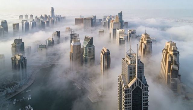 Fog-cloaked skyscrapers rising above marina and canal at dawn, dramatic waterfront skyline