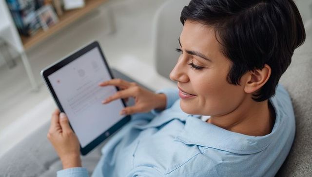 Reclining Woman Reading Tablet Wearing Blue Pajama Top on Sofa in Sunlit Living Room