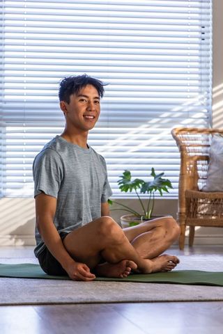Smiling asian man practicing seated meditation on yoga mat in sunlit living room